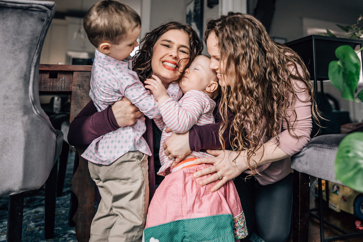 A same sex couple hugs and kisses their little boy and little girl during an in home photo session with Minneapolis family photographer, Kate Simpson.