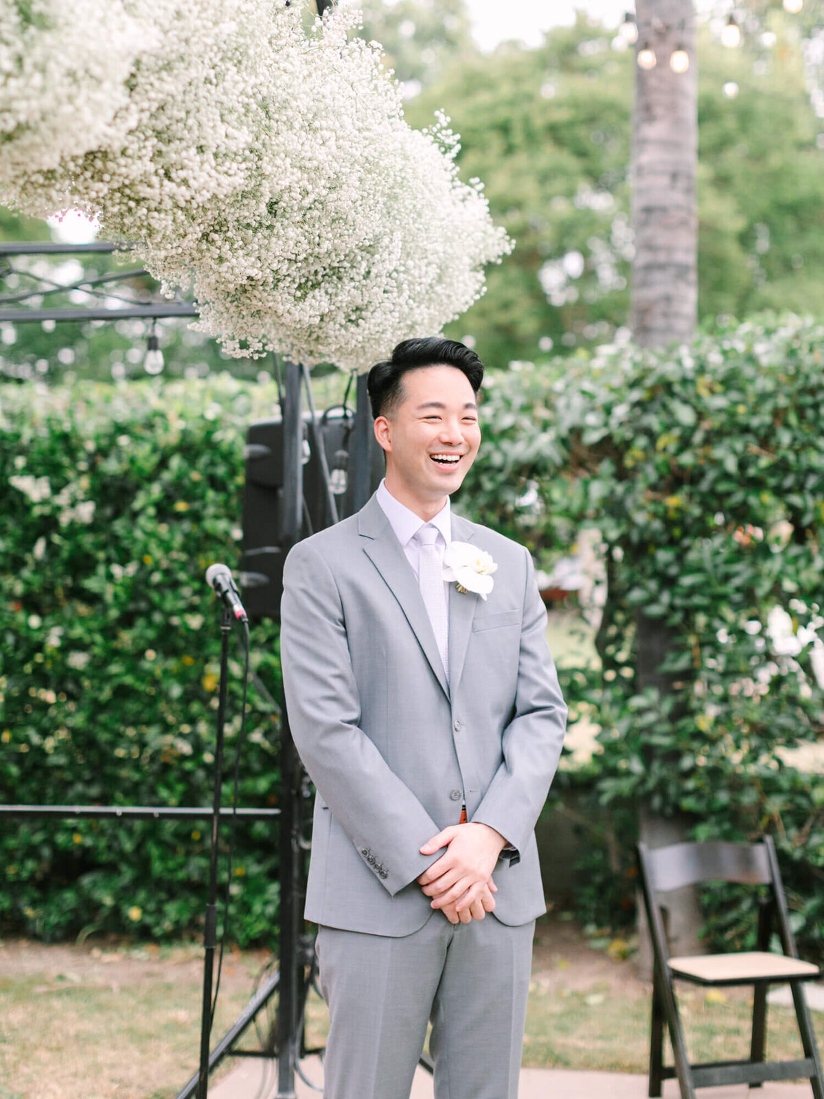 A joyful groom in a light gray suit smiles under a canopy of baby's breath flowers at Muckenthaler Mansion wedding venue. He's outdoors, with green foliage and a microphone stand nearby.