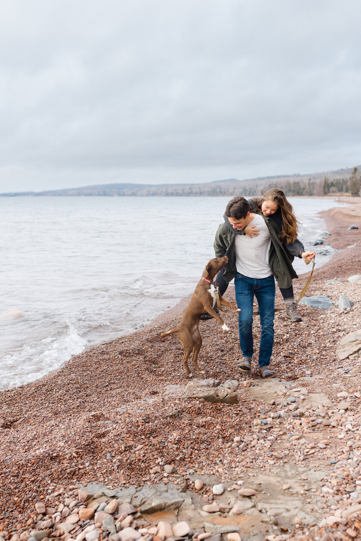 A married couple and their dog stroll on the shore of Lake Superior on a breezy day with a Duluth wedding photographer.