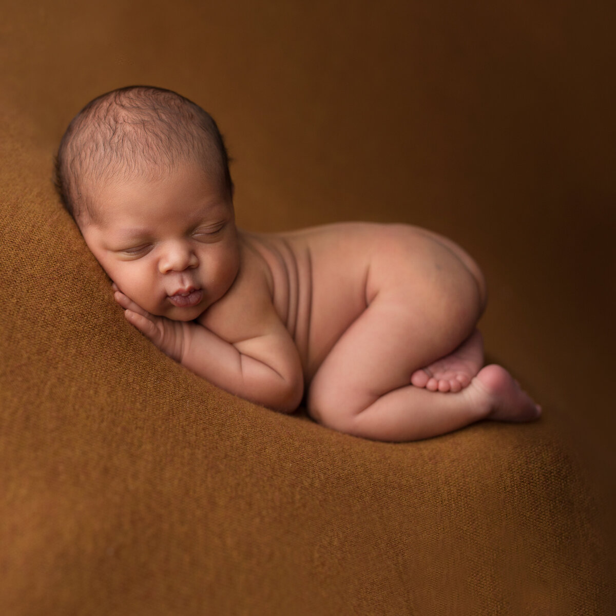 Black baby boy lies on his stomach with his feet crossed under him.  He is on a brown backdrop.  He has baby rolls and chubby cheeks.