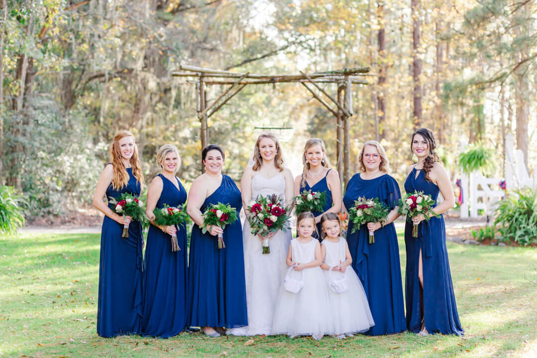 Bridesmaids in blue dresses stand with the bride and flowergirls in white dresses under a wooden arbor outdoors in a garden