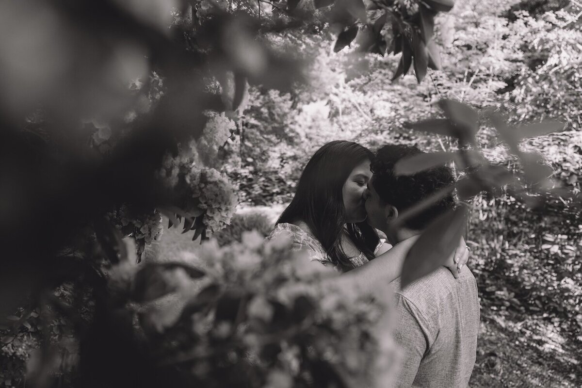 Black-and-white image of couple sharing a kiss through blurred leaves.