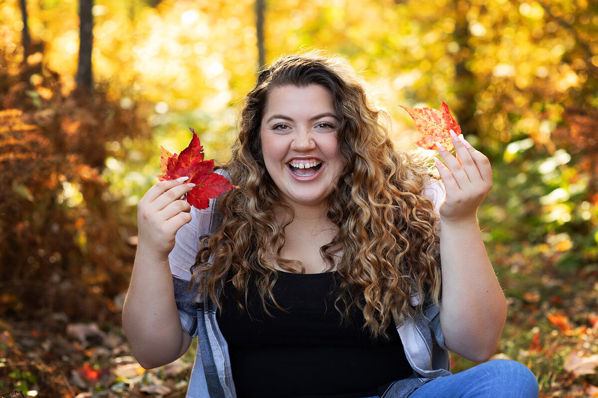 Studio 64 Photography captures happy portrait of girl sitting on ground holding fall colored leaves.