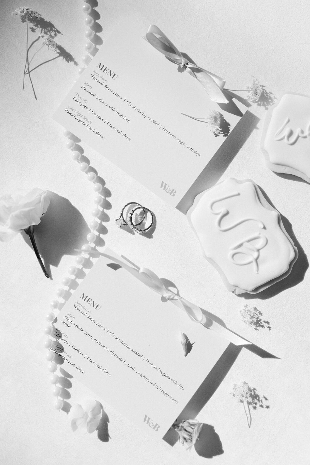 Black and white wedding flat lay featuring a custom printed menu card with satin ribbon, pearl string, monogrammed cookie, and wedding rings