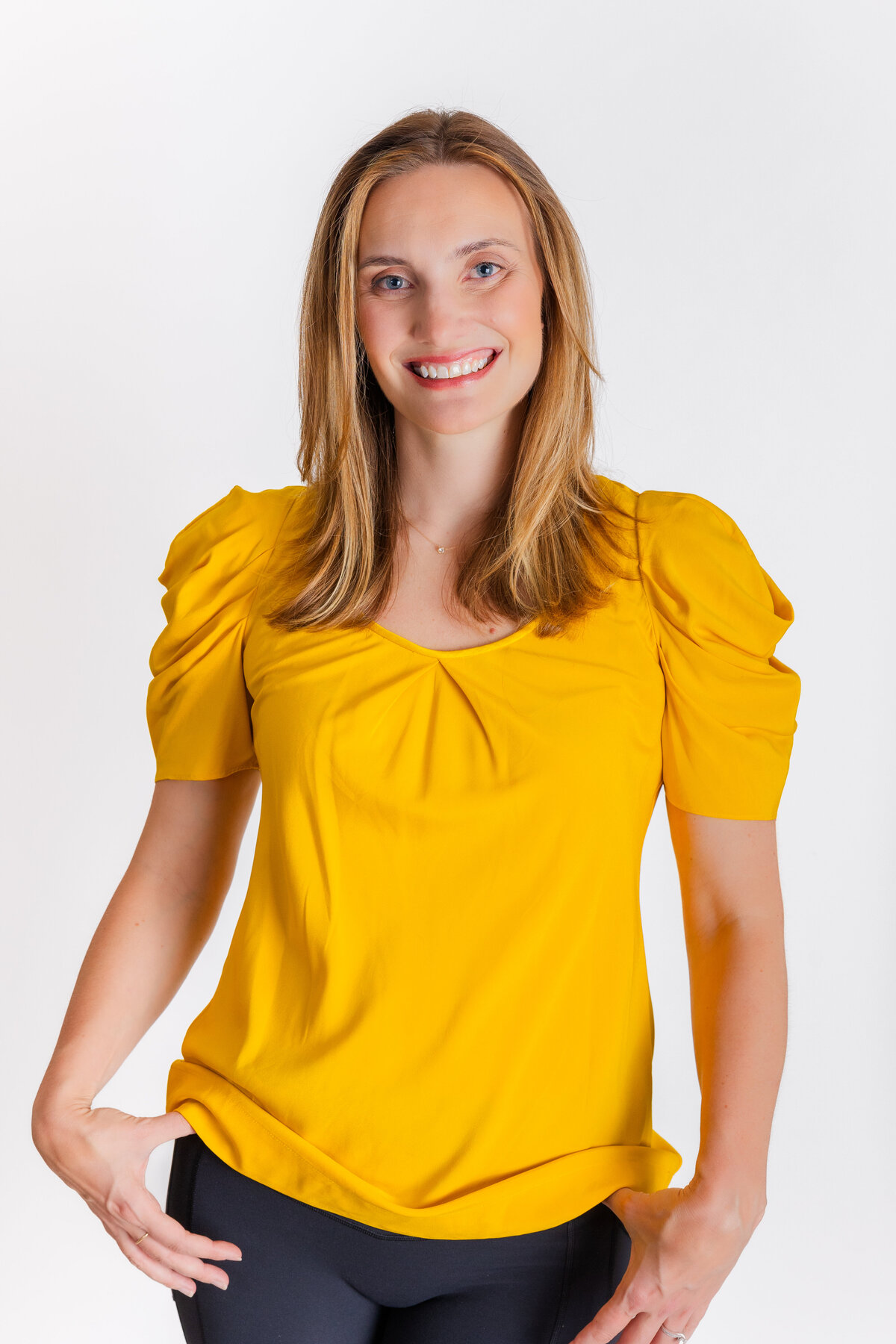 woman wearing dark jeans amd a yellow blouse stanfind hands in pockets in front of a white backdrop during portraits session