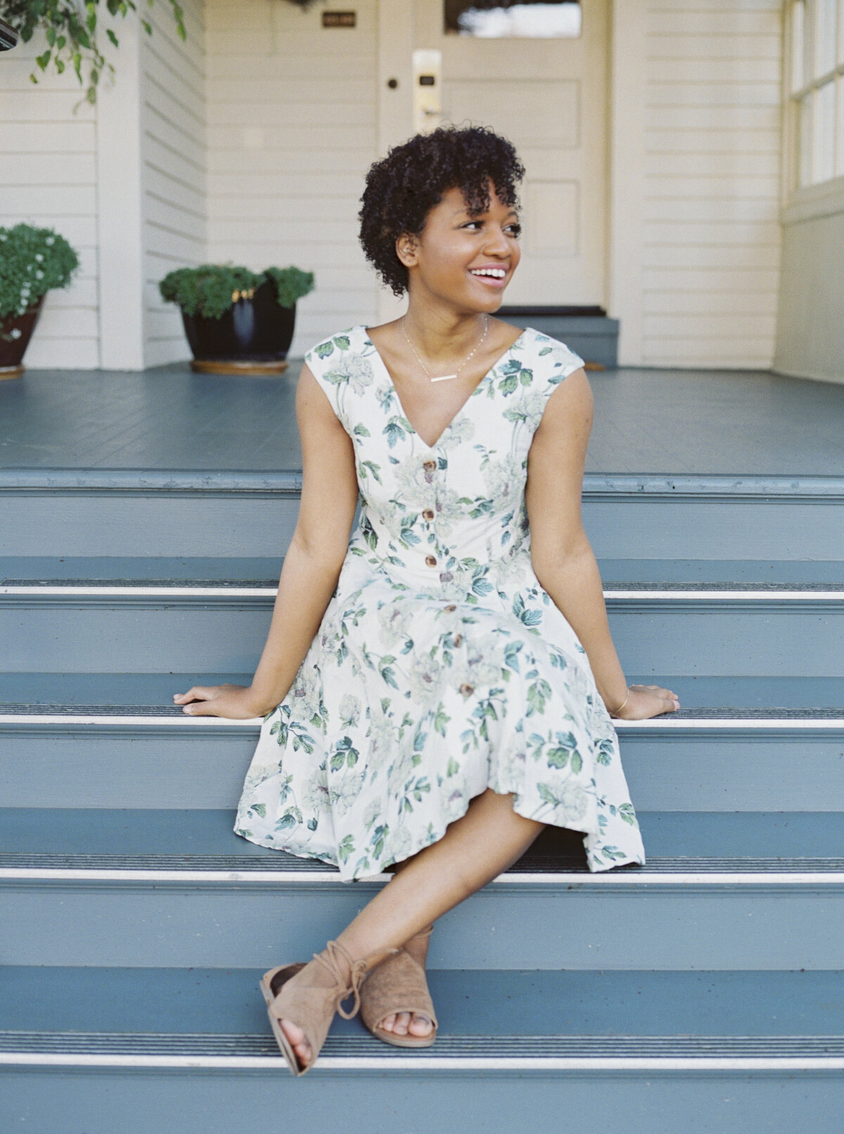 Black woman in floral dress smiling while seated on porch steps during NYC brand photo session.