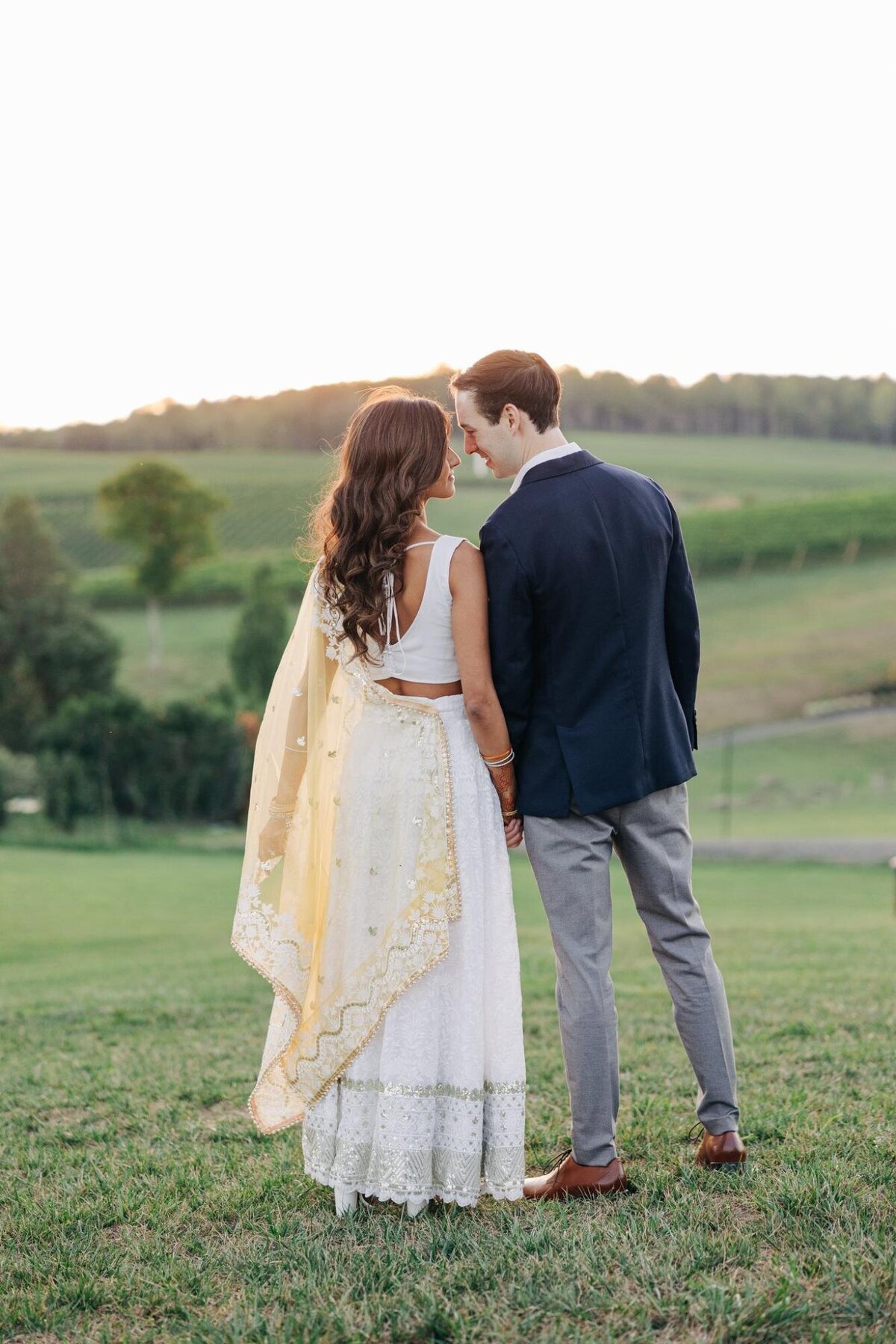 A bride and groom stand close together in a field, the bride in a white lace dress and the groom in a blue suit, facing a sunset.