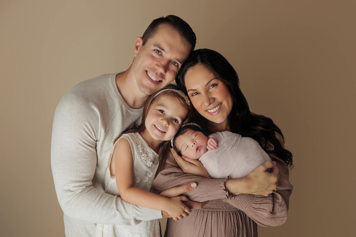 A happy family portrait featuring a father, mother, young daughter, and newborn baby. The mother holds the swaddled newborn while the daughter and father lovingly embrace them, all smiling at the camera.