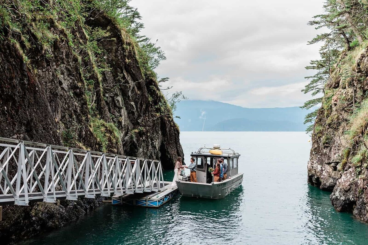 A bride walked down a dock to meet her groom and their guests in the flat bottomed water taxi that will take them to their vows location during their Kenai Fjords Alaska ELopement .  The boat is at the end of a sloping long metal dock running along the edge of the fjord cliff, covered with green moss.  A similar rocky cliff is viewable on the right side of the frame, with low mountains and clouds in the distance behind them across the water. 