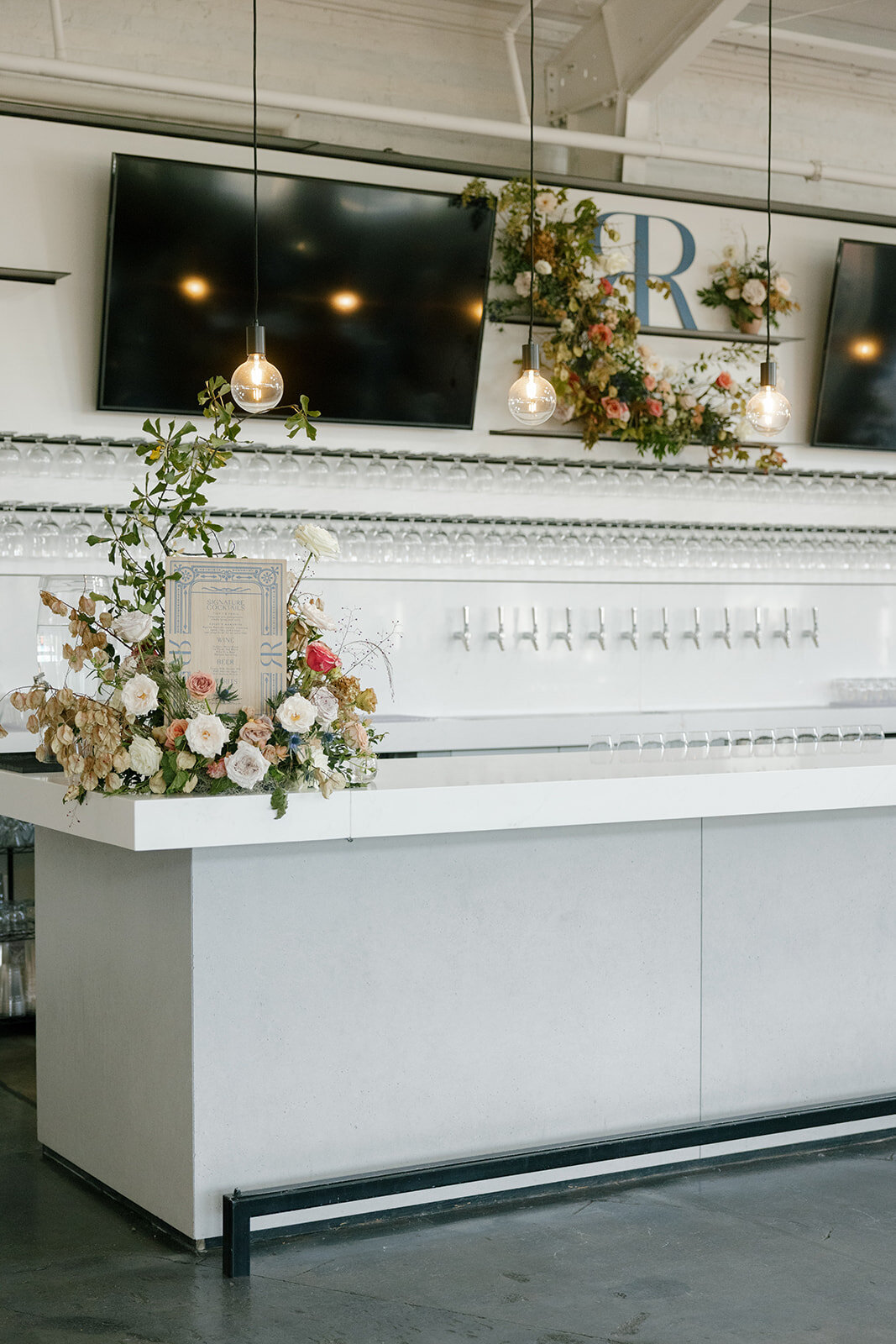 Lush floral install surrounds the bar menu in floral colors of taupe, mauve, dusty rose, and navy composed of petal heavy roses, raintree pods, thistle, dried hydrangea, and natural fall foliage. Design by Rosemary and Finch Floral Design at Raleigh, NC wedding.