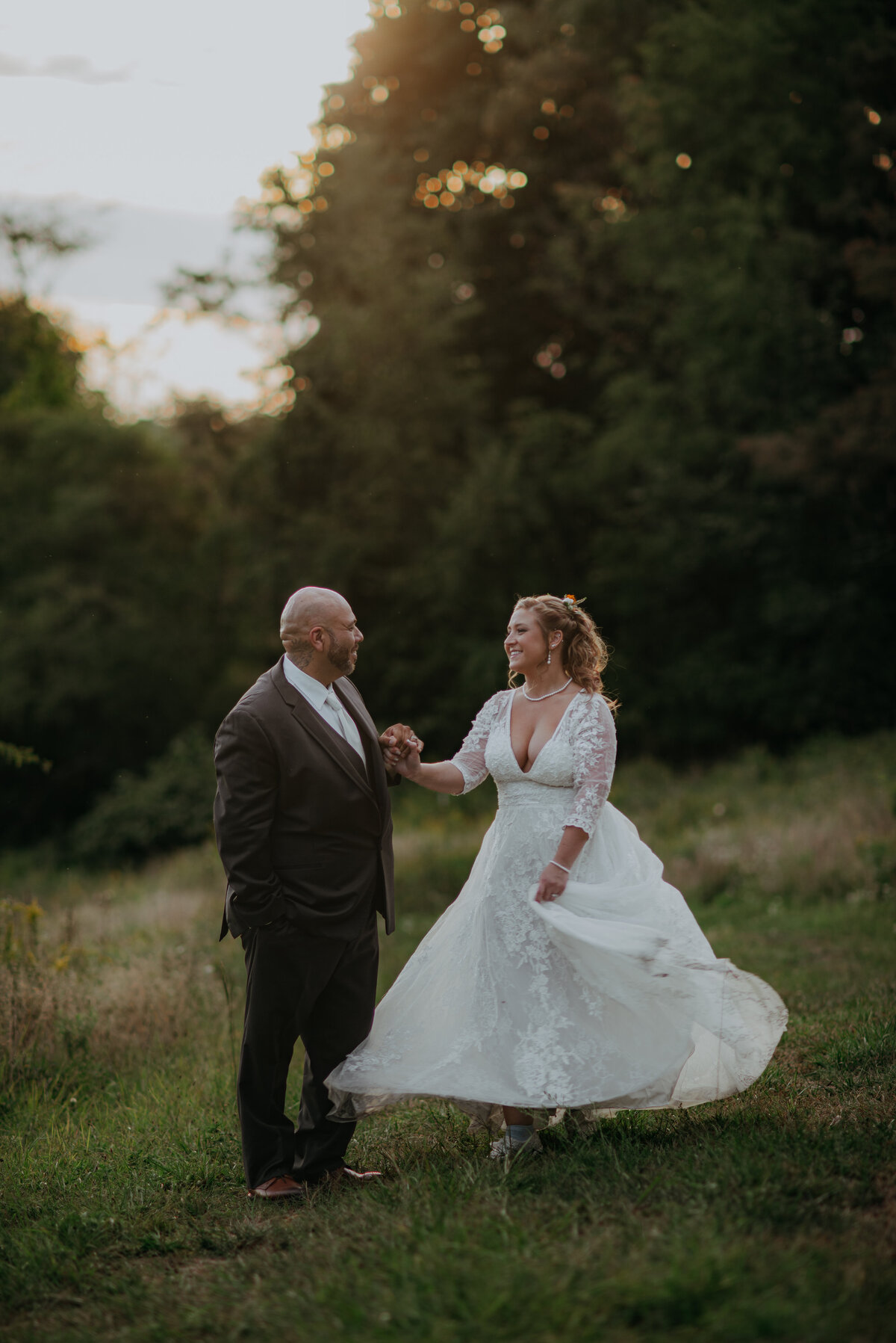 A groom holds the hand of his bride as she spins her dress in a moody woodsy golden hour full body portrait.