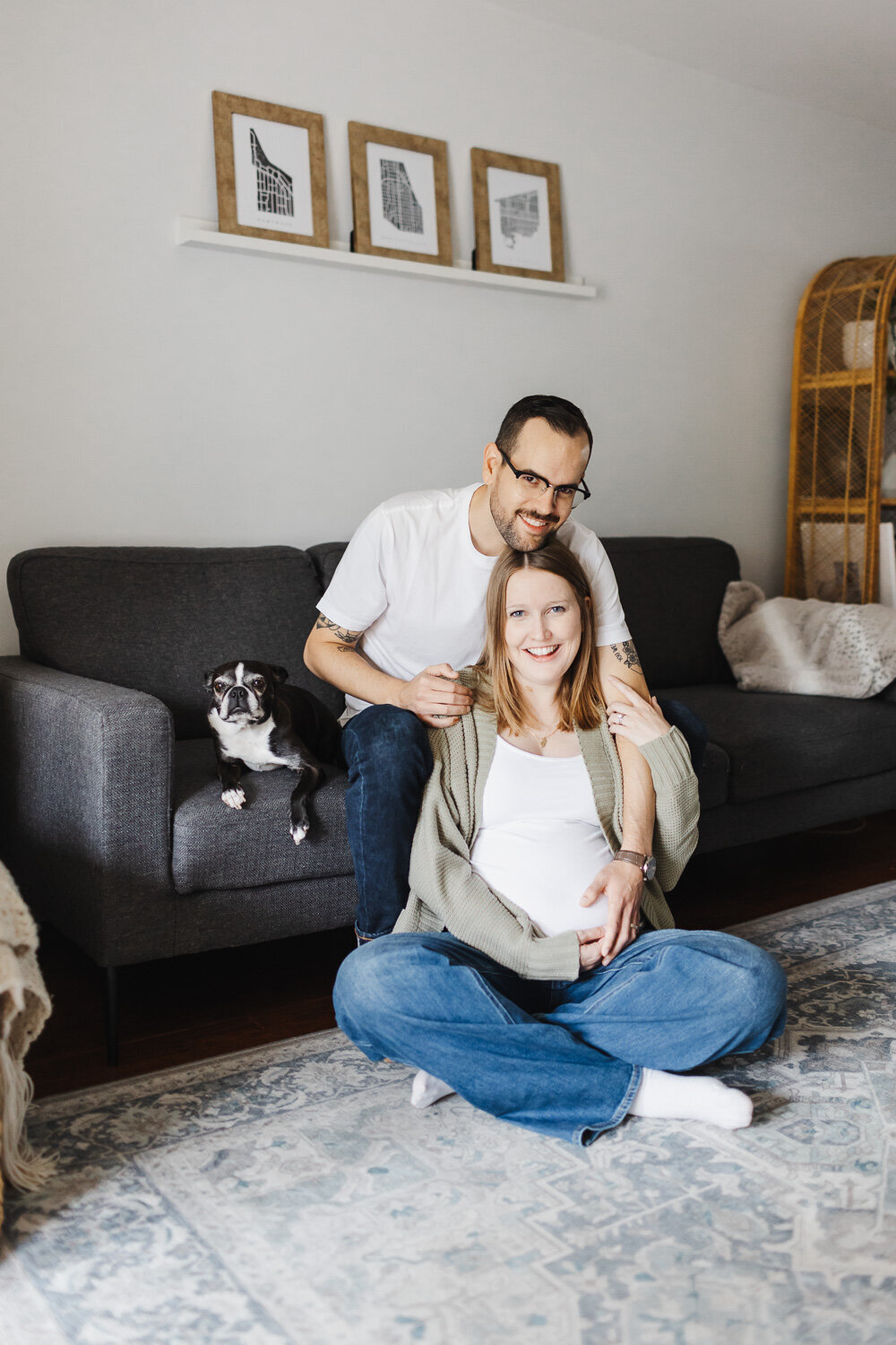 Expecting mom sitting on floor in front of couch. Husband is on the couch behind her and their pet dog is on the couch. All are smiling at the camera.