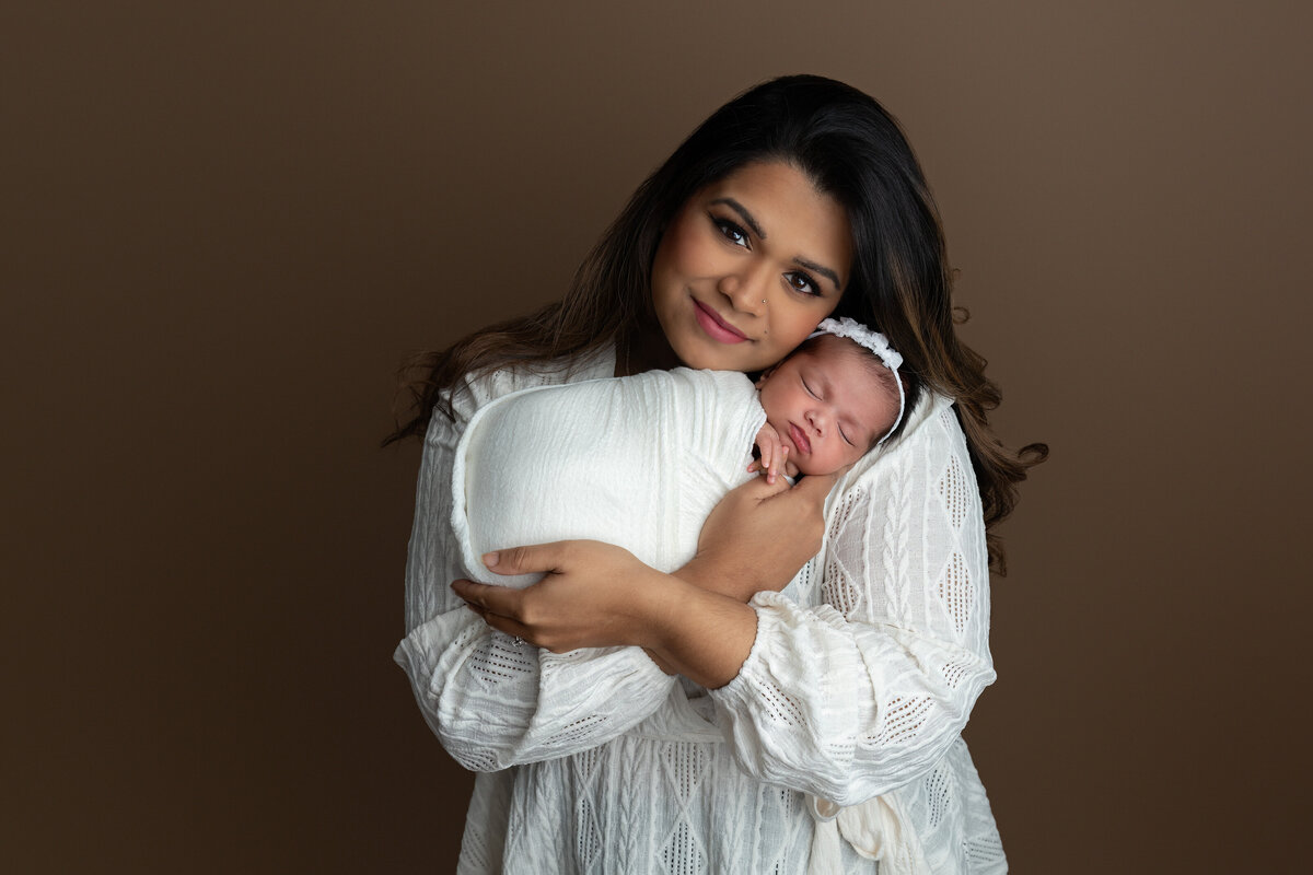An intimate mother and baby portrait taken in the studio, showing a mother holding her newborn daughter wrapped in soft white swaddling. Photographed at Meraki Photographic Creations in Paris, Ontario.