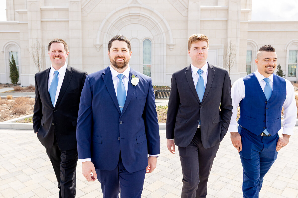  The groom and his groomsmen walk confidently toward the camera, all smiling and dressed in blue and black suits.