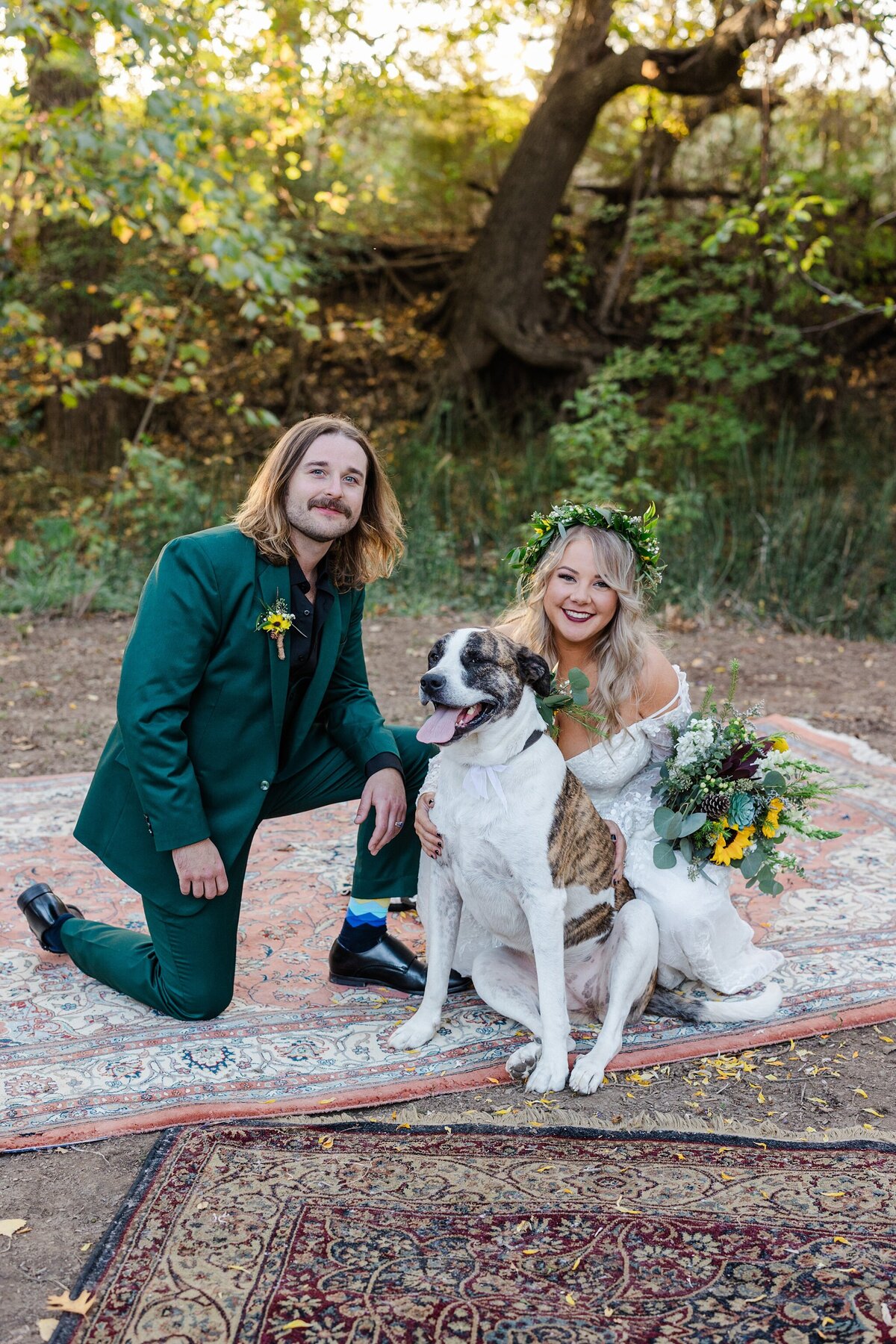A portrait of a bride, groom, and their dog on their wedding day in Fort Worth, Texas. The bride is on the right is wearing a long sleeve, intricate, white dress with a flower crown and a large bouquet. The groom is kneeling on the left and is wearing a green suit with a black dress shirt and boutonniere. Their dog is seated in front of the bride and is mostly white with large brown patches.