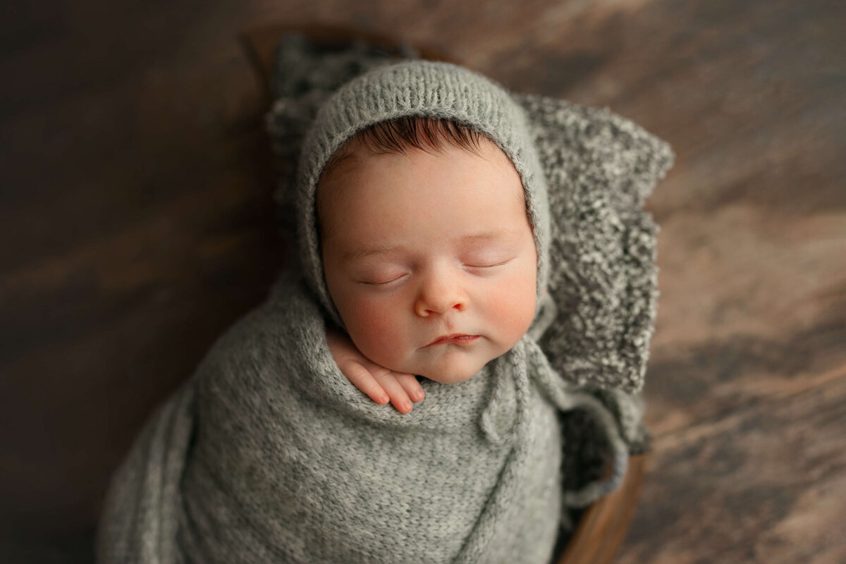 newborn baby wearing a grey wrap and bonnet laying on a pillow in a prop at a photography session