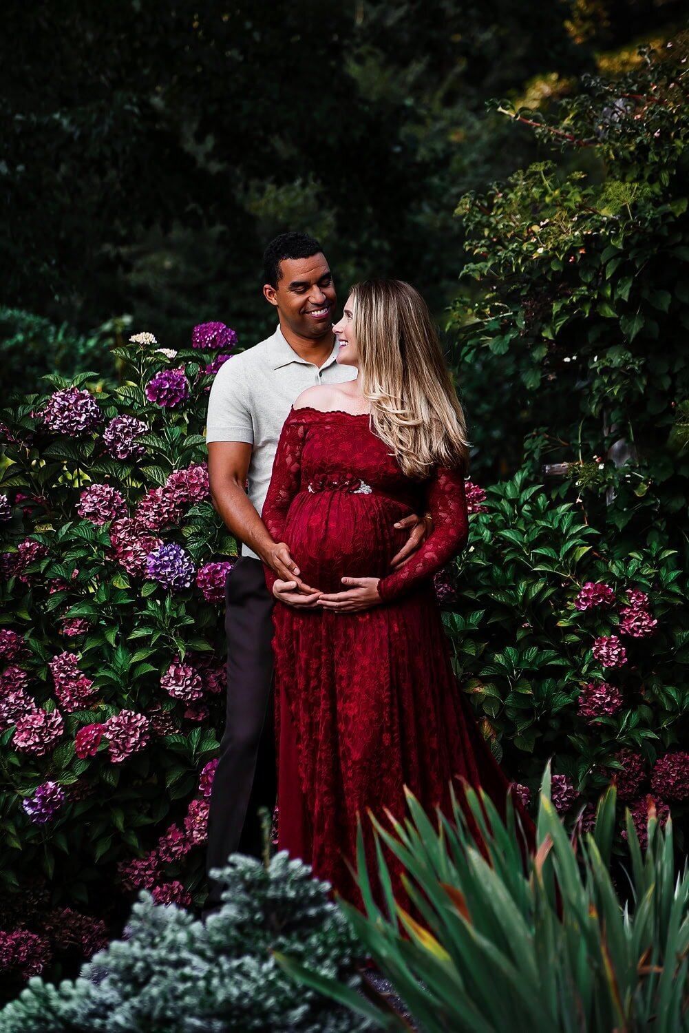 Expecting couple during an outdoor maternity session at Deer Lake Park in Burnaby, with the mother in a red lace dress surrounded by a blooming garden of pink flowers.