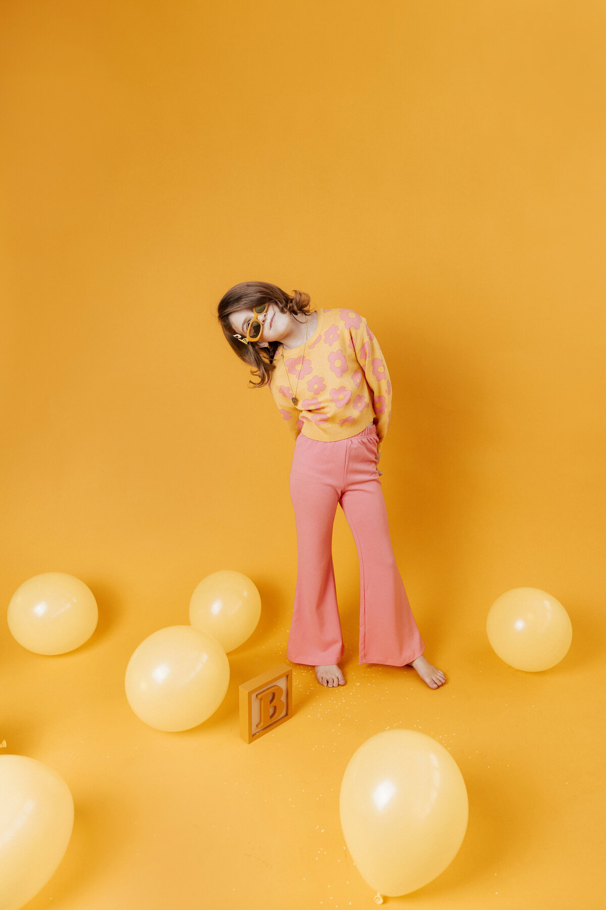 Playful retro-inspired child portrait in a bold yellow and pink studio setup, featuring a young girl in flared pants and flower print sweater surrounded by balloons and a wooden block with the letter B. Captured in Greenville, South Carolina by Dani Lacey Photographs.
