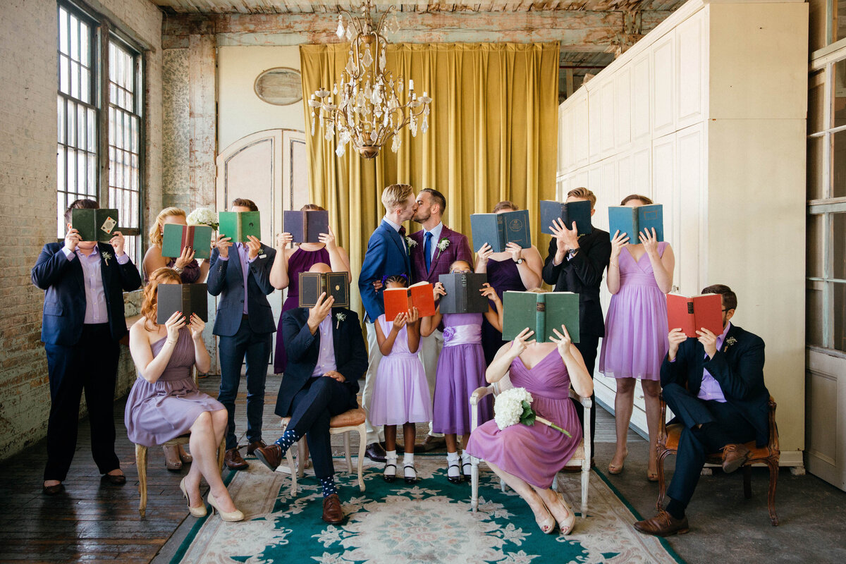 Wedding party holding books over their faces while two grooms kiss.