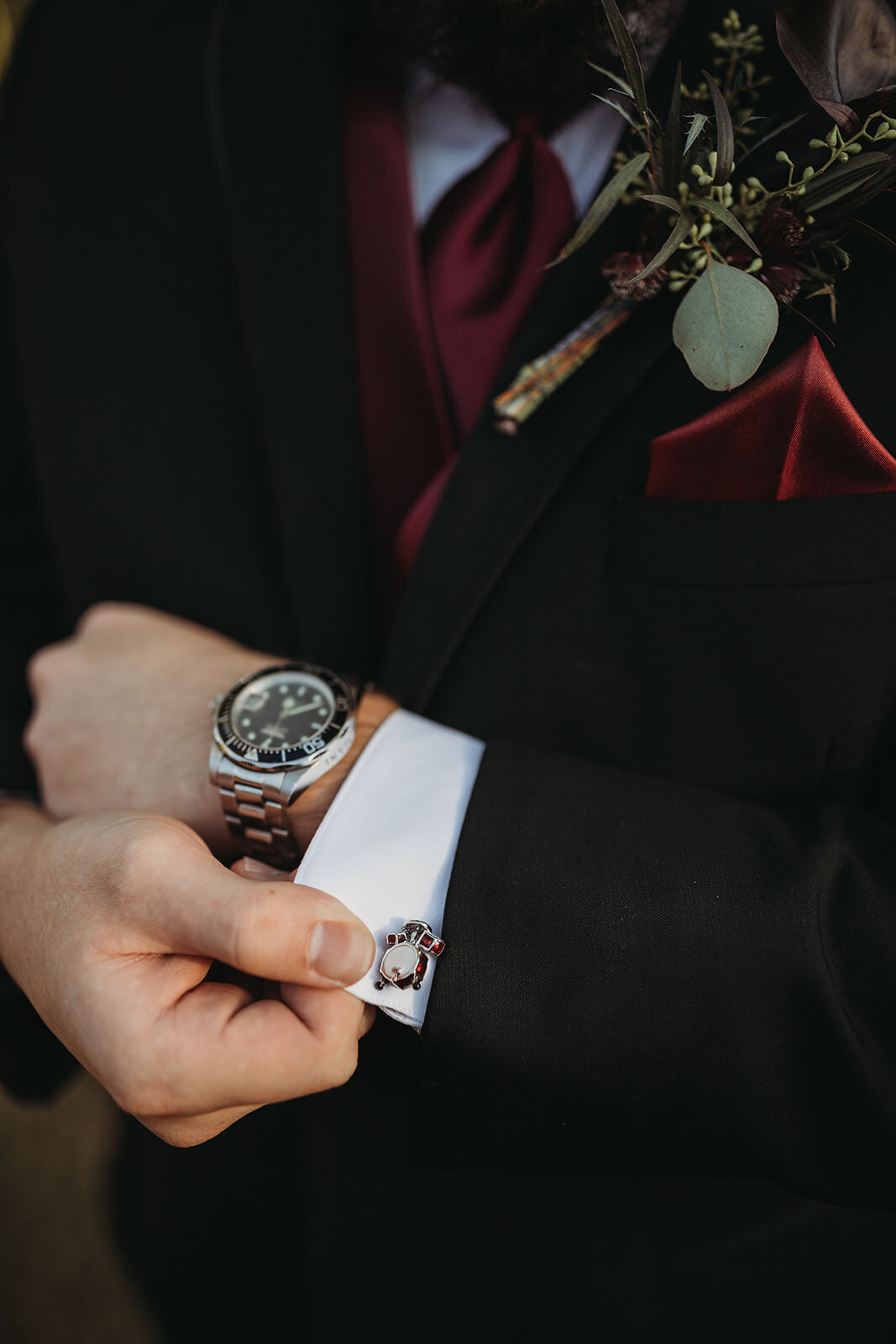 Detail of a groom's black suit, showing a drum set cufflink and a hand adjusting the cuff, with a maroon tie and boutonniere