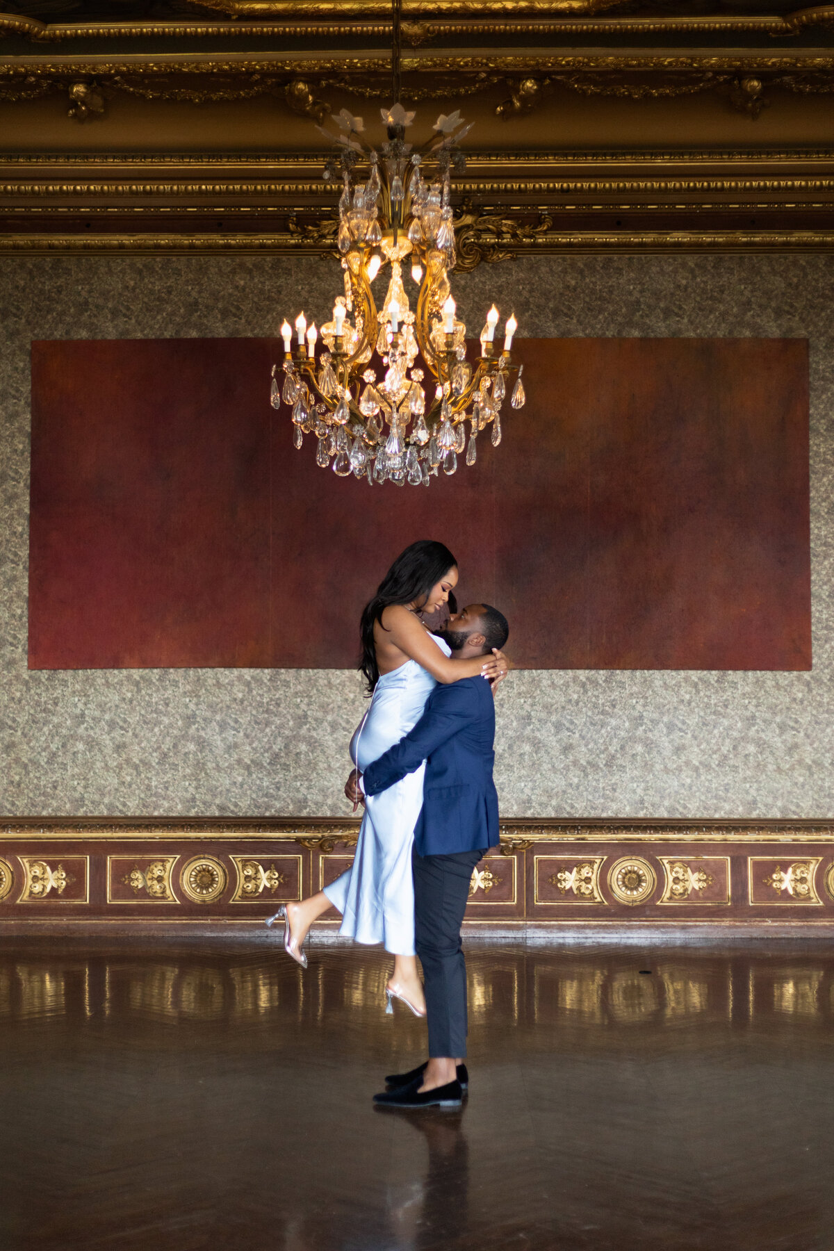 Ochre Court Mansion  | Kelsey Sheehan Photography Timeless Rhode Island Weddings | A couple embraces under an ornate chandelier in an elegant room. The woman, in a silver dress, is lifted by the man, creating a romantic and joyful atmosphere.
