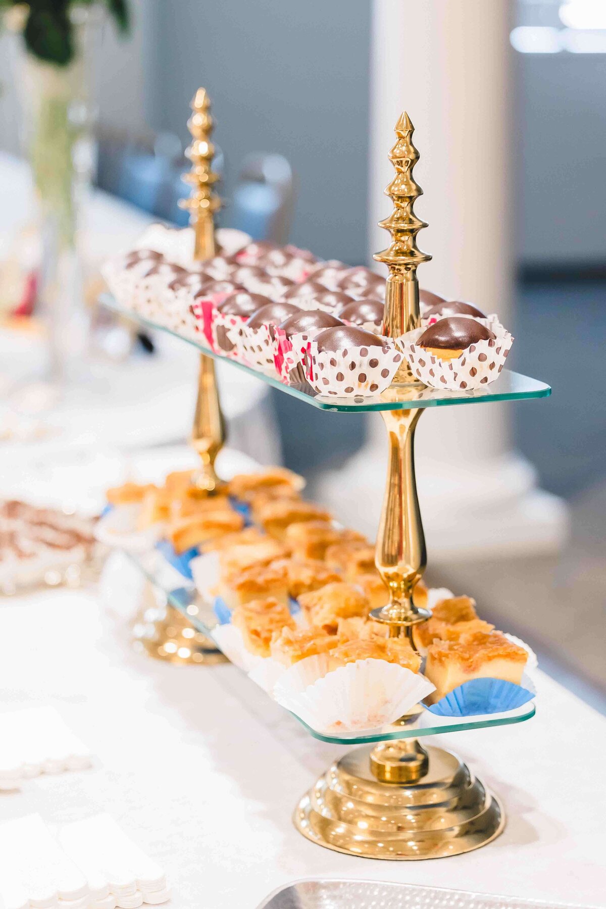 Two-tiered gold stand displaying assorted sweets, captured by an event photographer in Fayetteville, NC. The top tier has chocolate-covered treats with polka dot and floral liners. The bottom tier holds small pastry squares in paper cups. The stand is set on a white tablecloth.