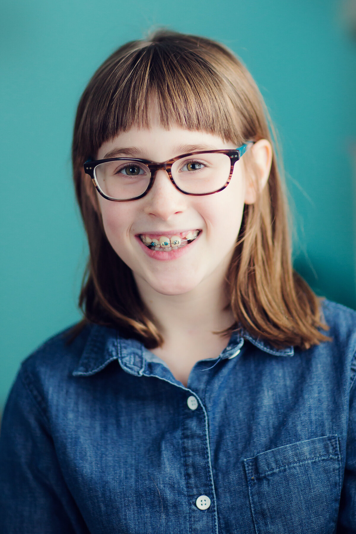 Portrait of girl smiling at the camera with glasses, braces, and a denim shirt.