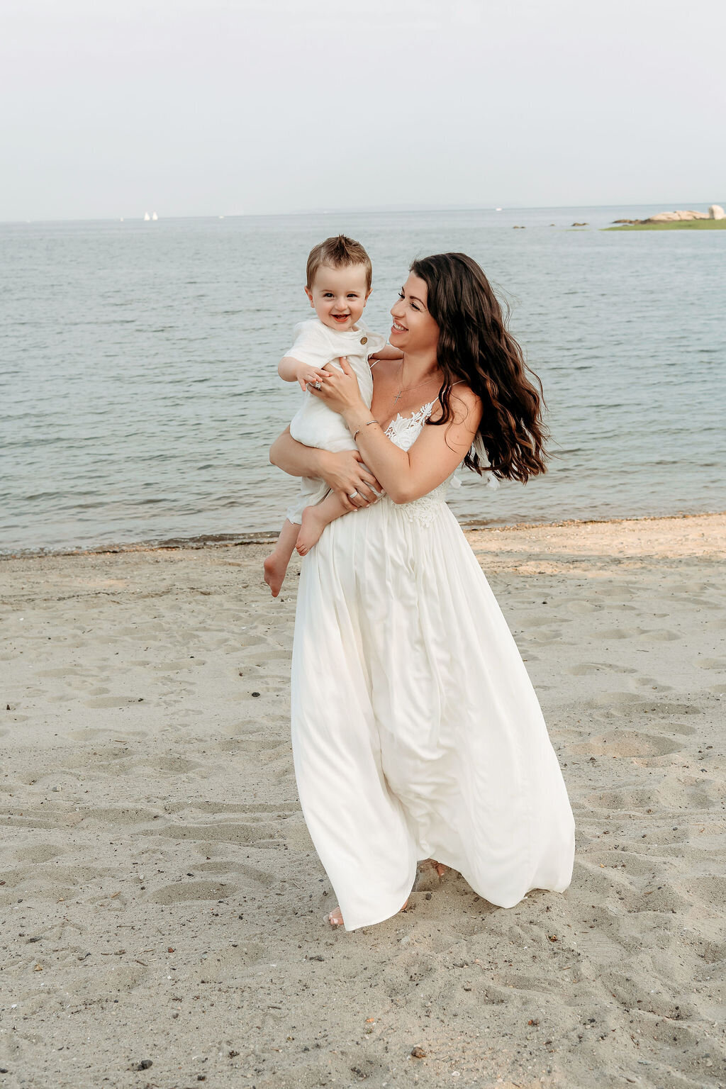 Pregnant mother in white flowing gown holds smiling toddler at waterfront