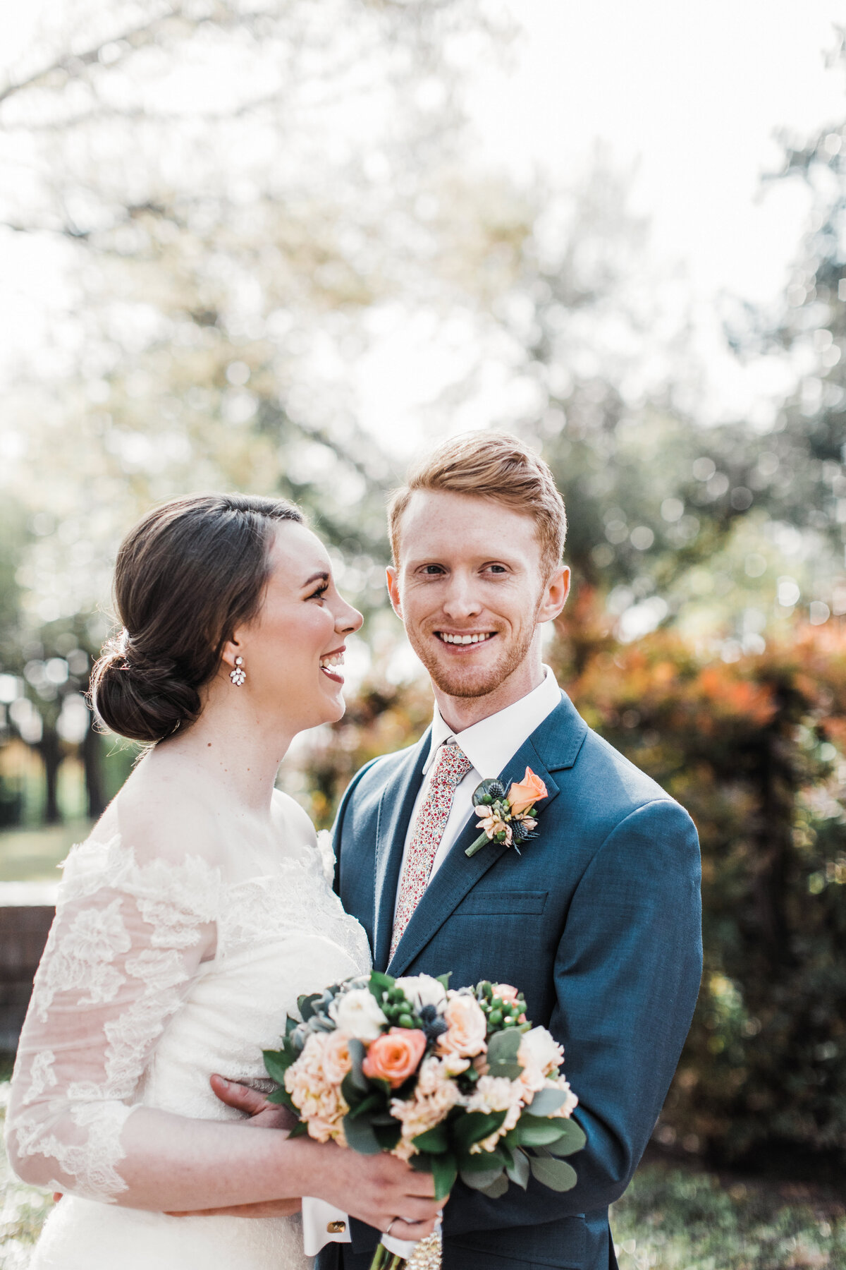 Portrait of a bride and groom outside of the Marty Leonard Community Chapel on their wedding day in Fort Worth, Texas. The groom smiles toward the camera while holding the bride close who is smiling joyfully at him. The bride is on the left and is wearing a long sleeve, intricate, white dress while holding a bouquet. The groom is on the right and is wearing a navy suit with a floral tie and boutonniere.