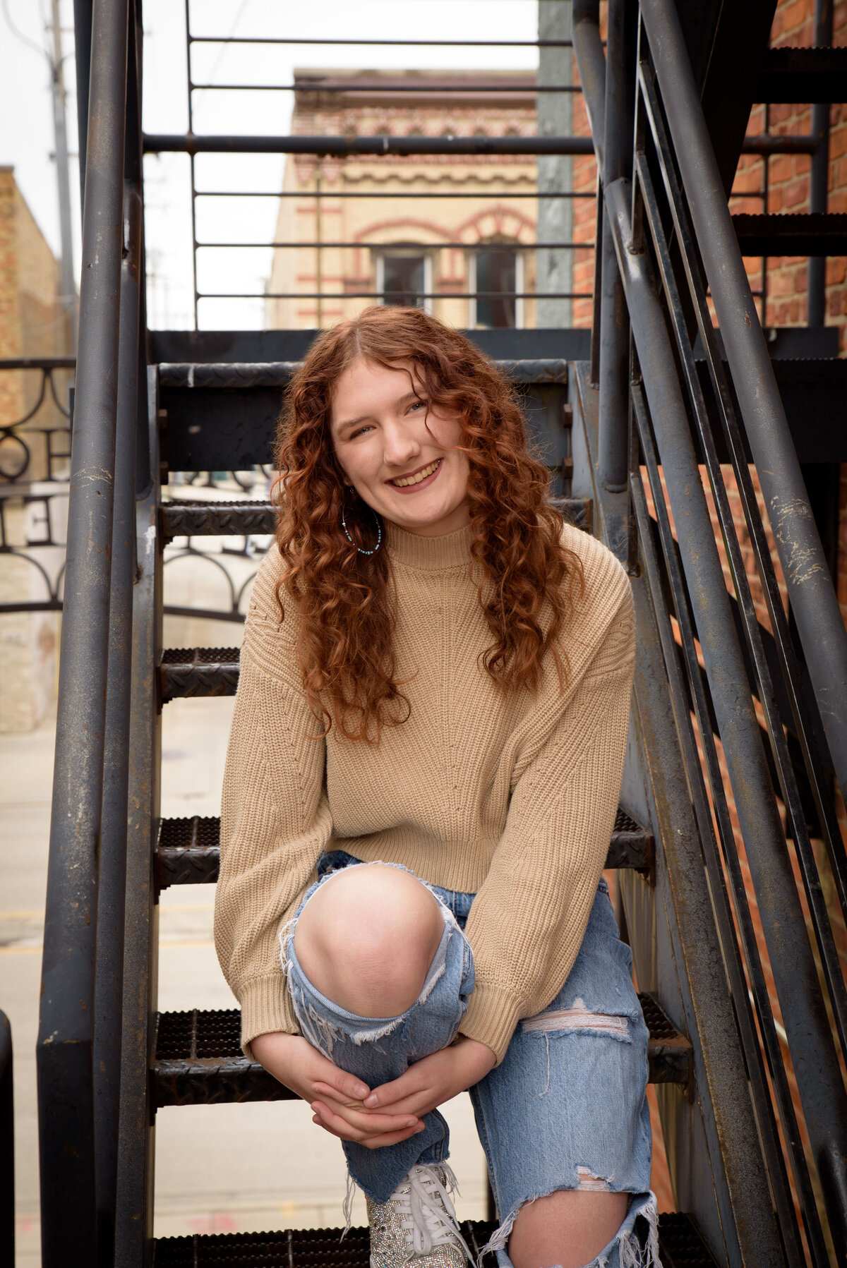 Green Bay East High School senior girl sitting on metal stairs wearing a tan sweater and jeans in urban setting in downtown Green Bay, Wisconsin.