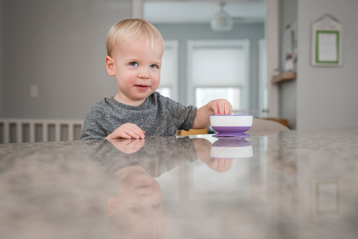 A toddler sits at a kitchen counter, smiling as he reaches for something in a small bowl.