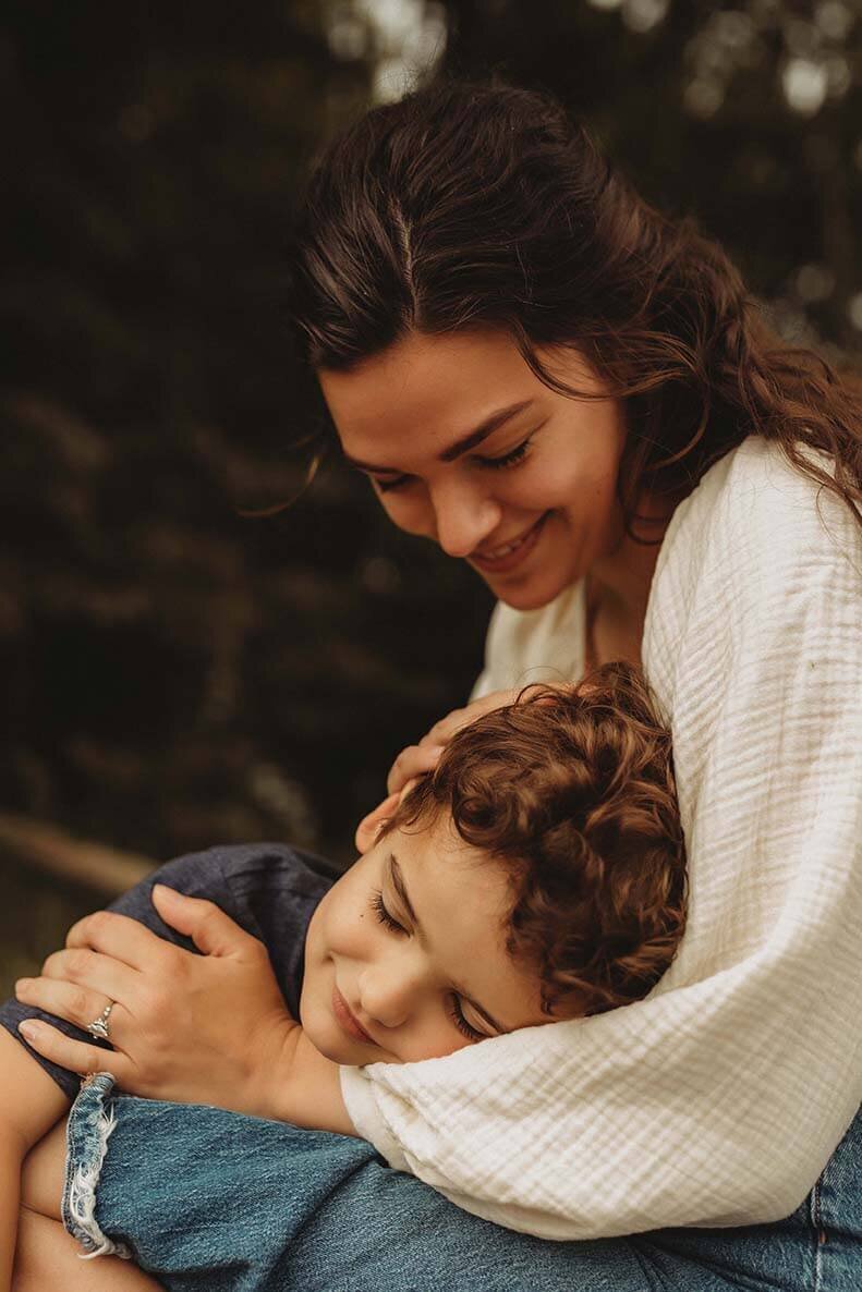 a son hugs his mothers lap with his eyes closed while his mom gently plays with his hair