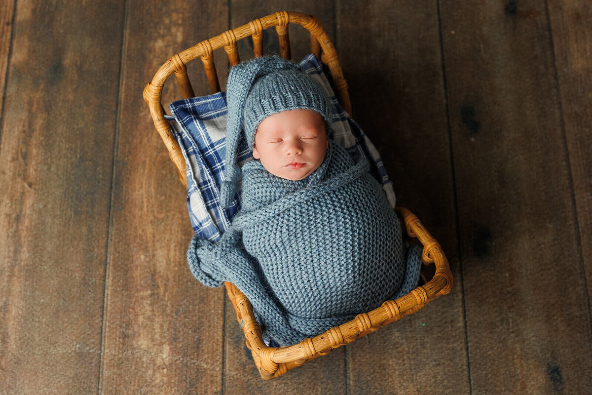 A peaceful newborn, swaddled in a soft blue knitted blanket with a matching hat, lies in a woven basket lined with a plaid fabric. The setting features rustic wooden flooring that adds warmth to the serene scene.