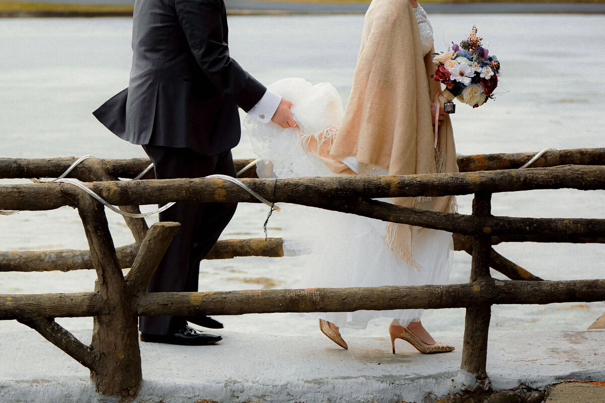 A bride in a white dress and shawl holds a bouquet while her groom helps lift her dress as they cross a rustic wooden fence near the water—beautifully captured by a film photographer NJ.