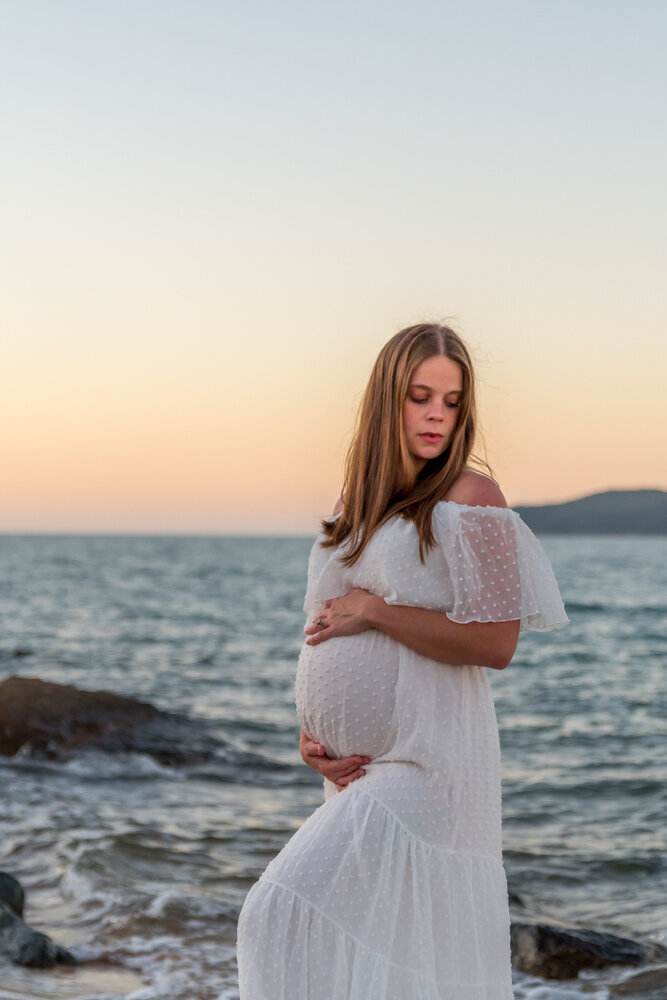 pregnant woman in white gown holding baby belly at the beach at sunset - Townsville Maternity Photography by Jamie Simmons