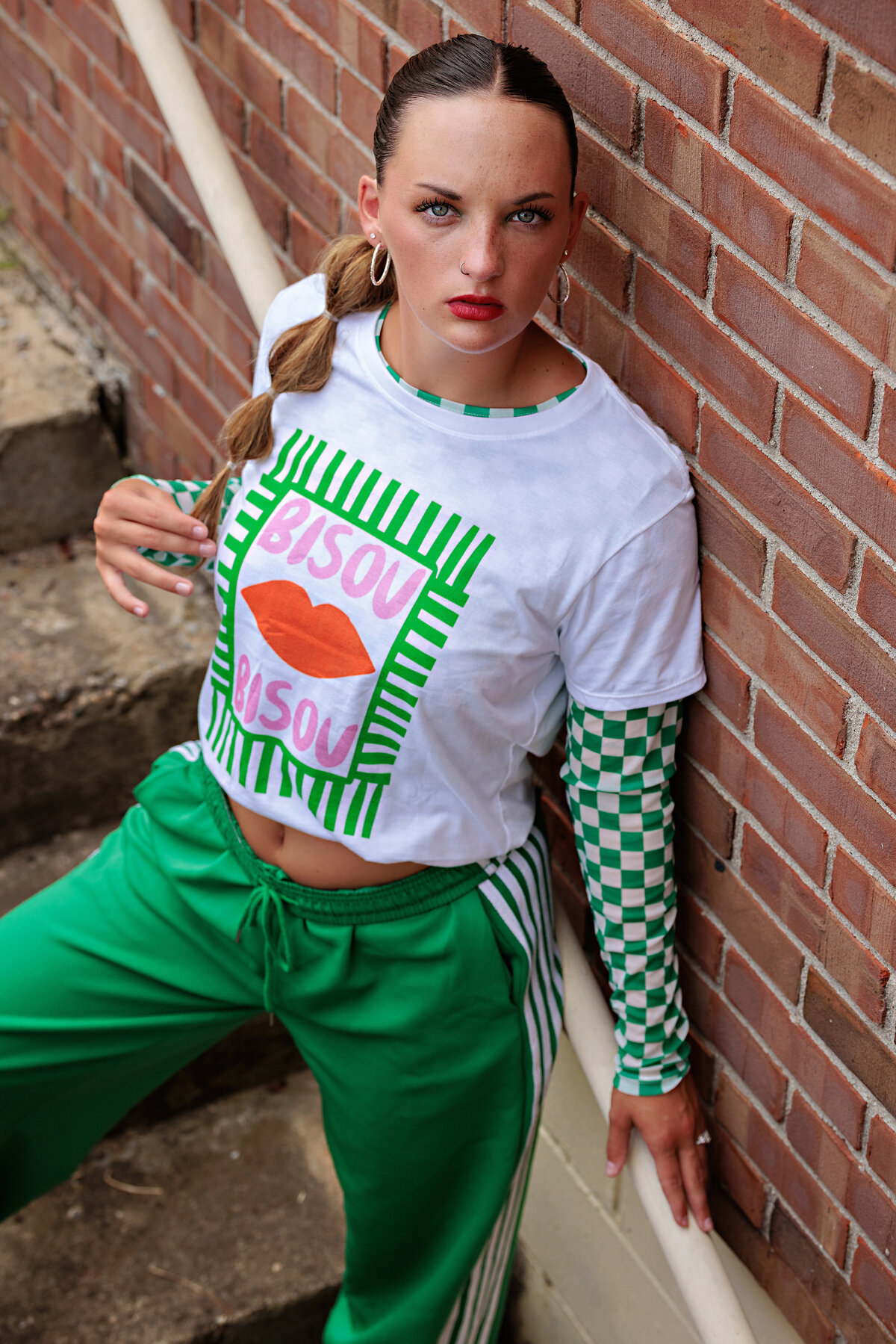 Stylish high school senior portrait featuring a teen in an emerald green outfit with checkered long sleeves layered under a graphic tee, green jogger pants, and a distinctive ponytail hairstyle, posed on a concrete staircase against a brick wall in Charlotte NC.