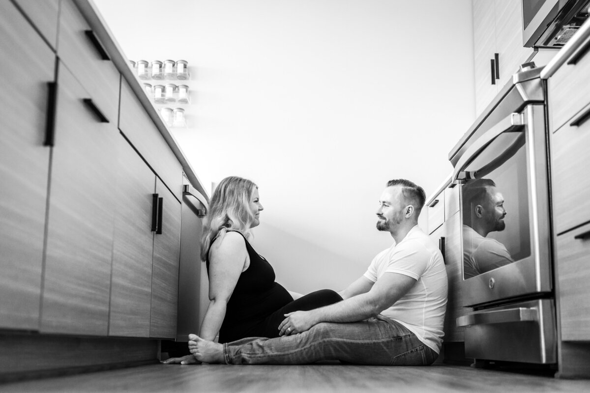 A couple sits on the floor of a modern kitchen, facing each other and holding hands, sharing a quiet and intimate moment together.