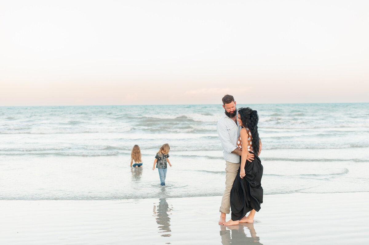 Parents hugging each other on the beach at sunset with the kids playing in the ocean in the background