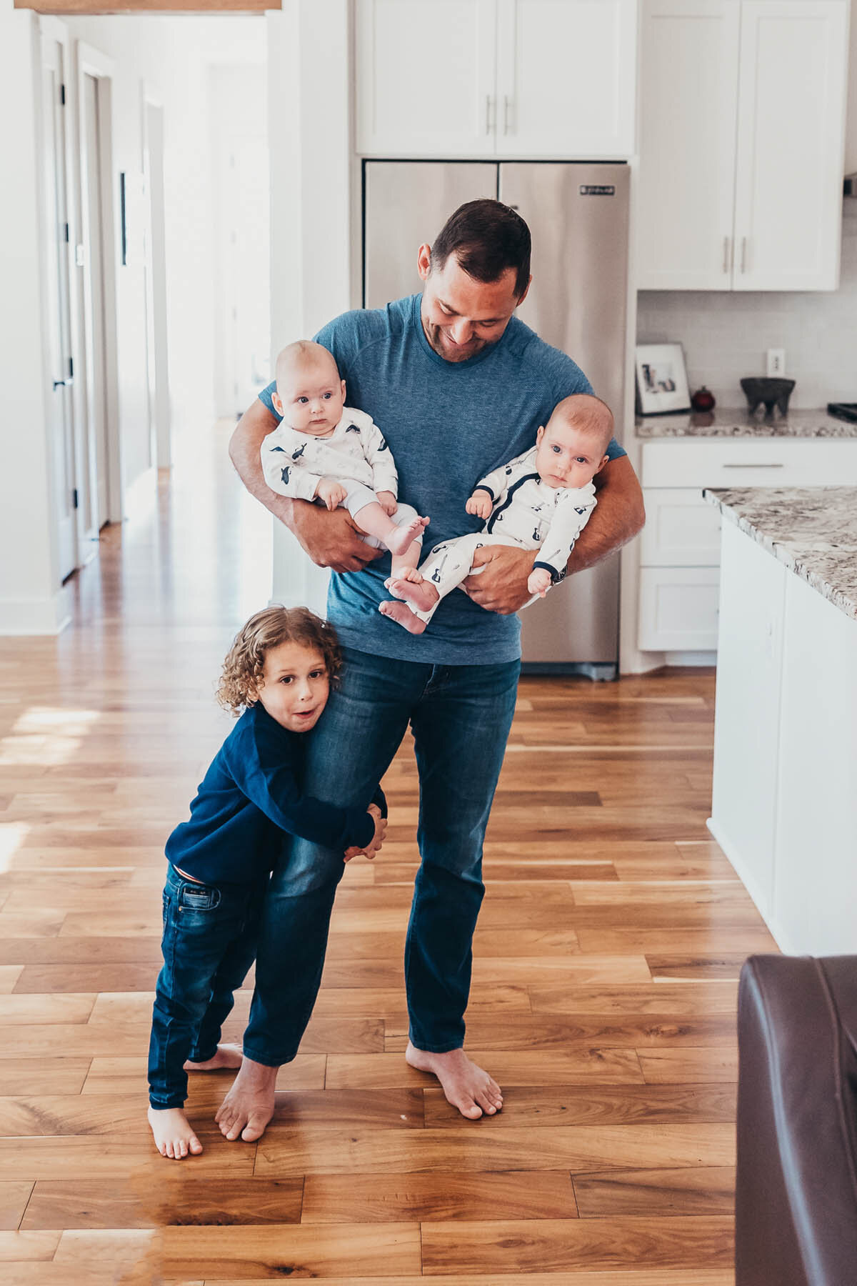 a father holds a baby in each arm while his toddler hugs his leg