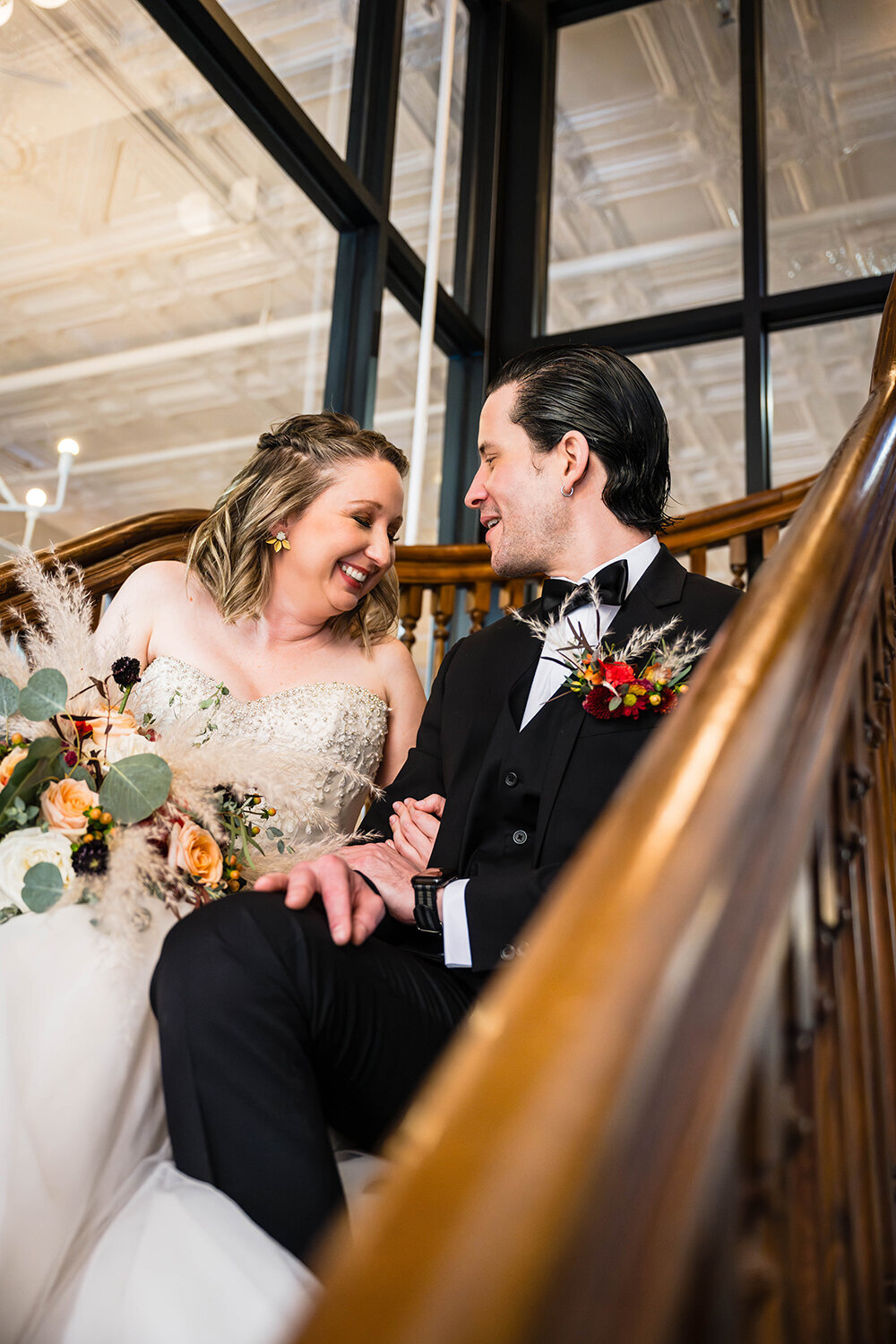 A couple on their elopement day sit side-by-side on a stairwell in the renovated Fire Station One hotel in Roanoke, Virginia and smile and laugh at one another.