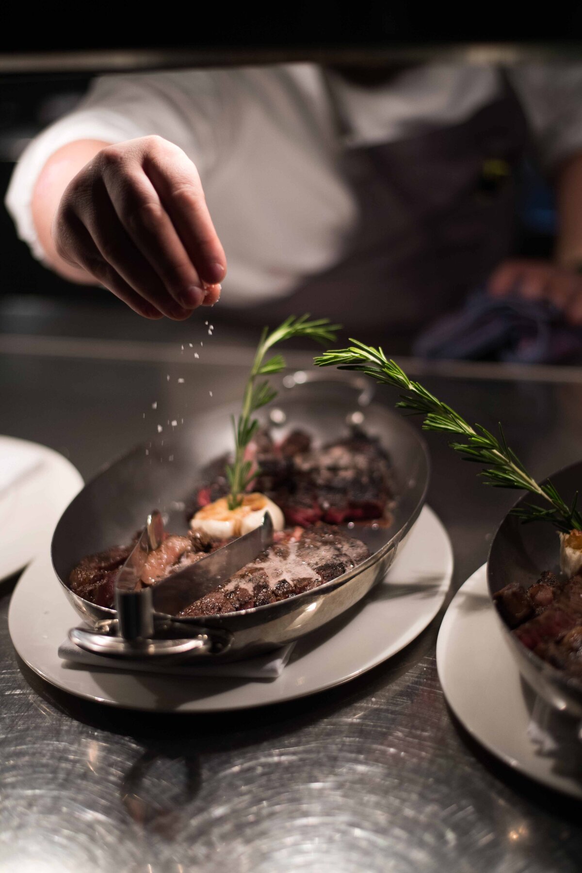 An image of a chef's hand sprinkling salt on a steak with a sprig of rosemary