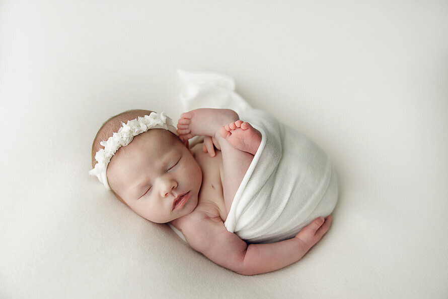 Newborn baby wrapped in white sleeps on a white backdrop.