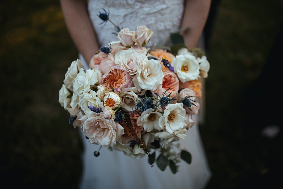 the bride wearing a lace dress holds her bouquet of peach, blush, ivory and white flowers with a small cascade of greenery