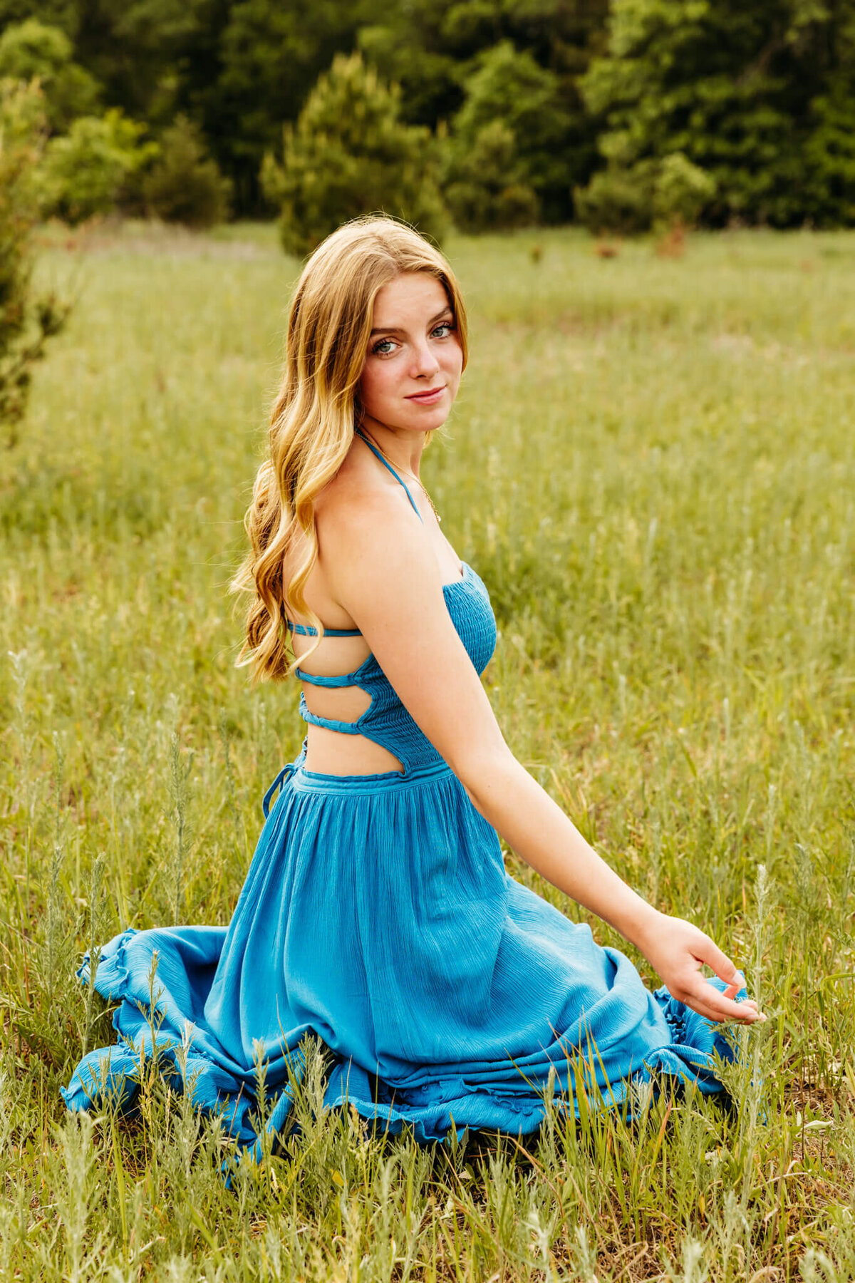 pretty high school girl in a strappy blue dress kneeling in the grass and looking back over shoulder