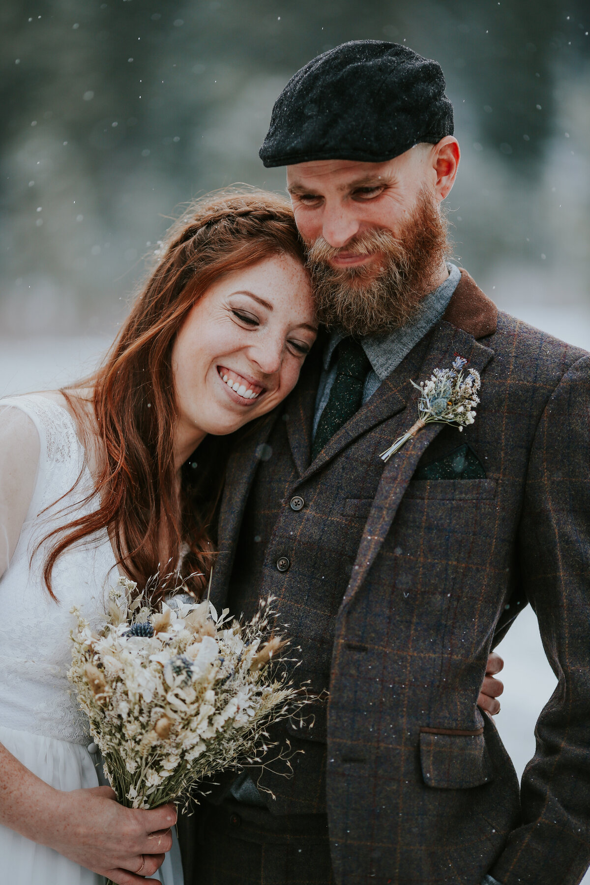 Smiling bride leans into grooms shoulder as snow gently falls.