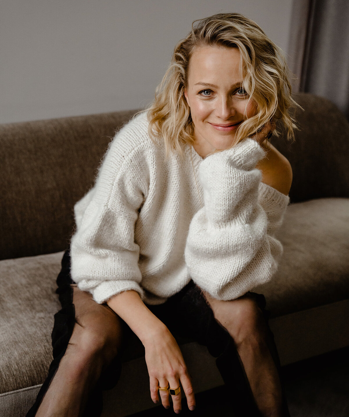 Portret van Tess Milne in een witte trui. Ze zit op een bank en kijkt lachend de camera in