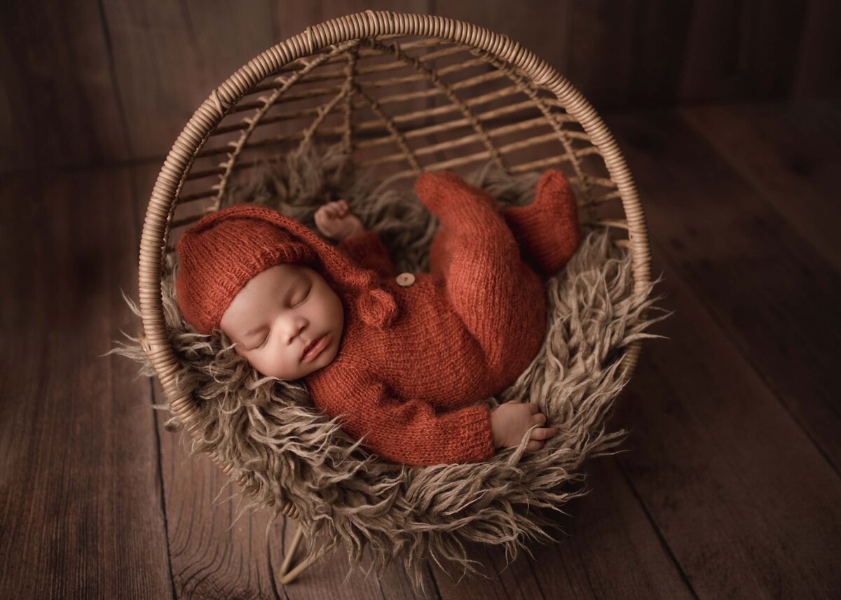 Baby boy in burnt orange knit romper with matching sleepy time hat sleeping in a boho style basket bed