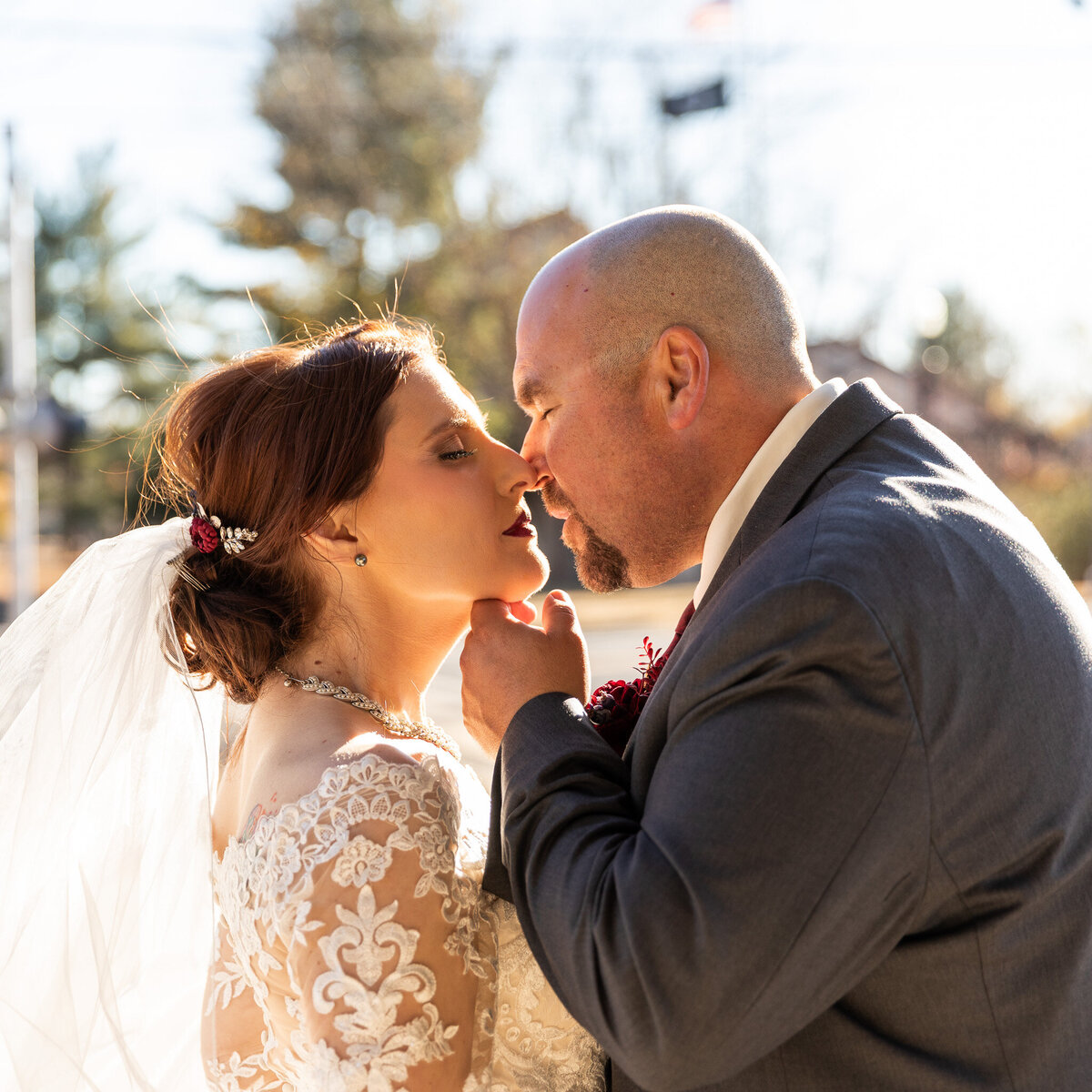 A groom gently lifts his bride’s chin as they lean in for a kiss, bathed in golden afternoon sunlight. The bride wears a lace gown with a flowing veil and jeweled hairpiece, while the groom in a charcoal suit draws close, capturing a tender and romantic moment.