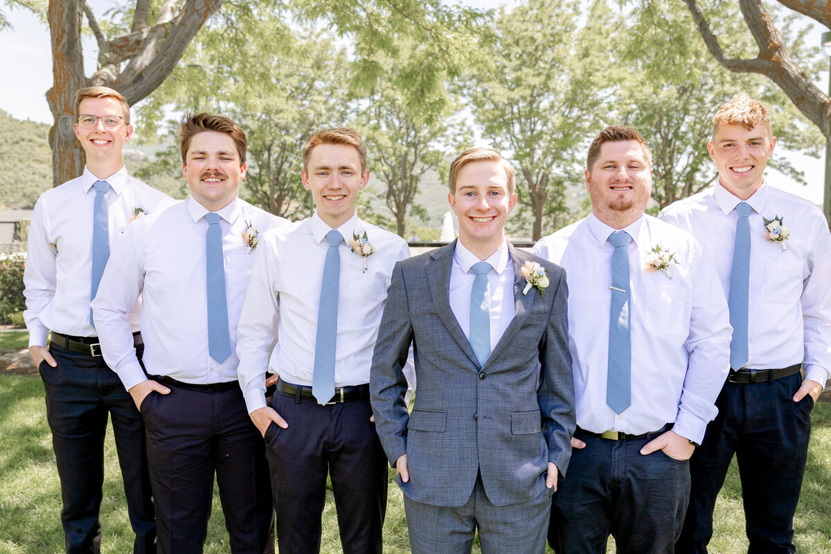 The groom and his groomsmen, all in crisp white shirts and blue ties, stand together in a sunny garden setting.