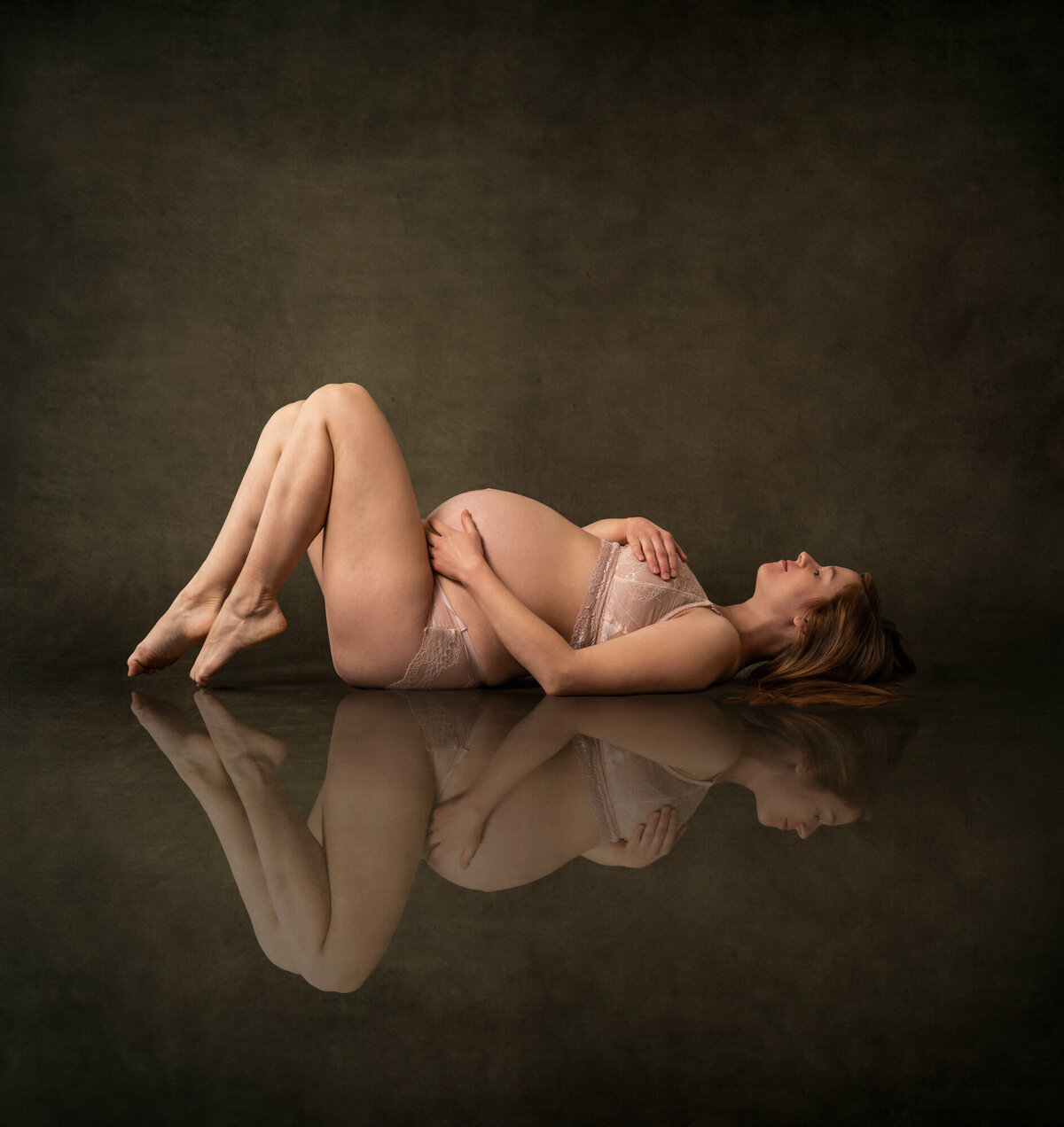 A beautiful mama to be in lingerie lays on the floor cradling her baby bump